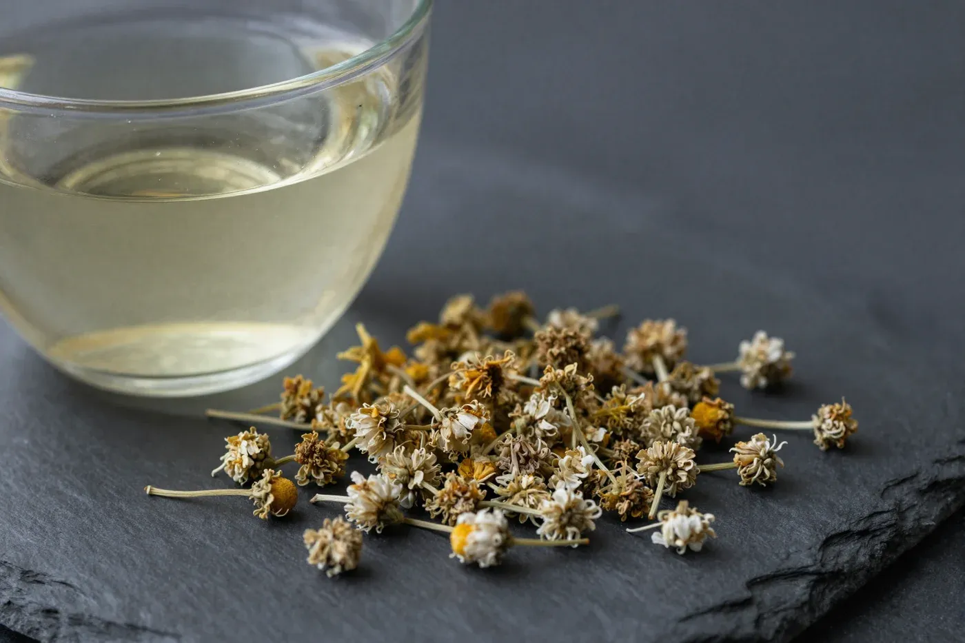 A glass cup of chamomile tea with chamomile flowers on a dark wooden surface.