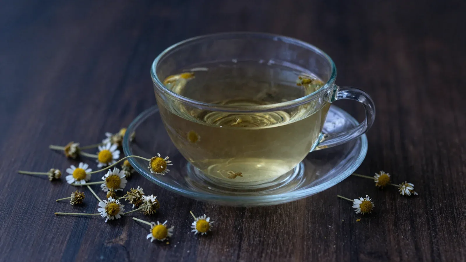 A cup of chamomile tea and dried chamomile flowers on a dark surface in dim evening light.