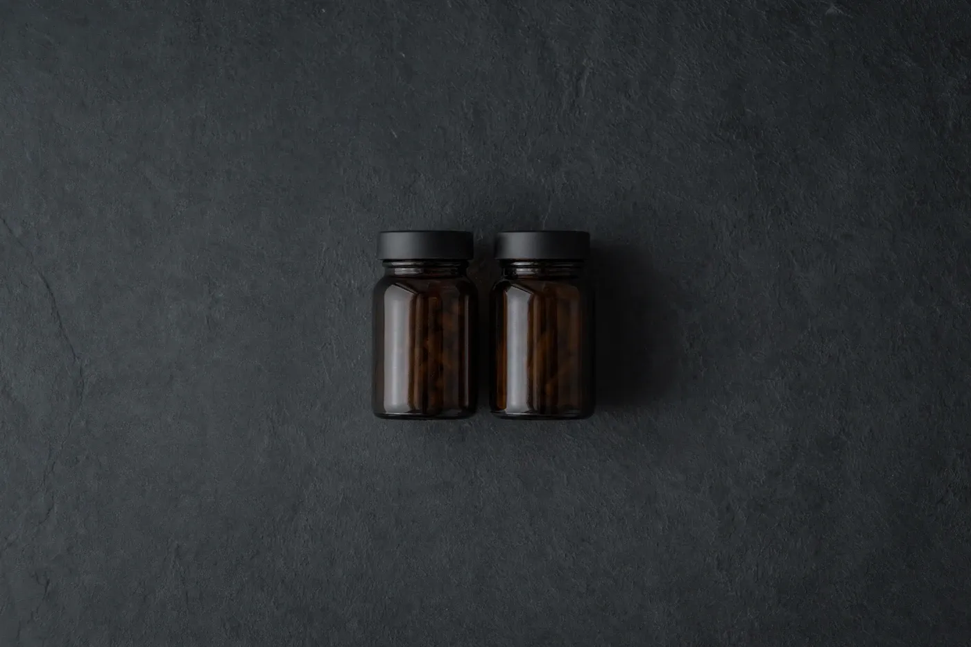 Two small unlabeled supplement bottles side by side on a dark slate surface in soft light.