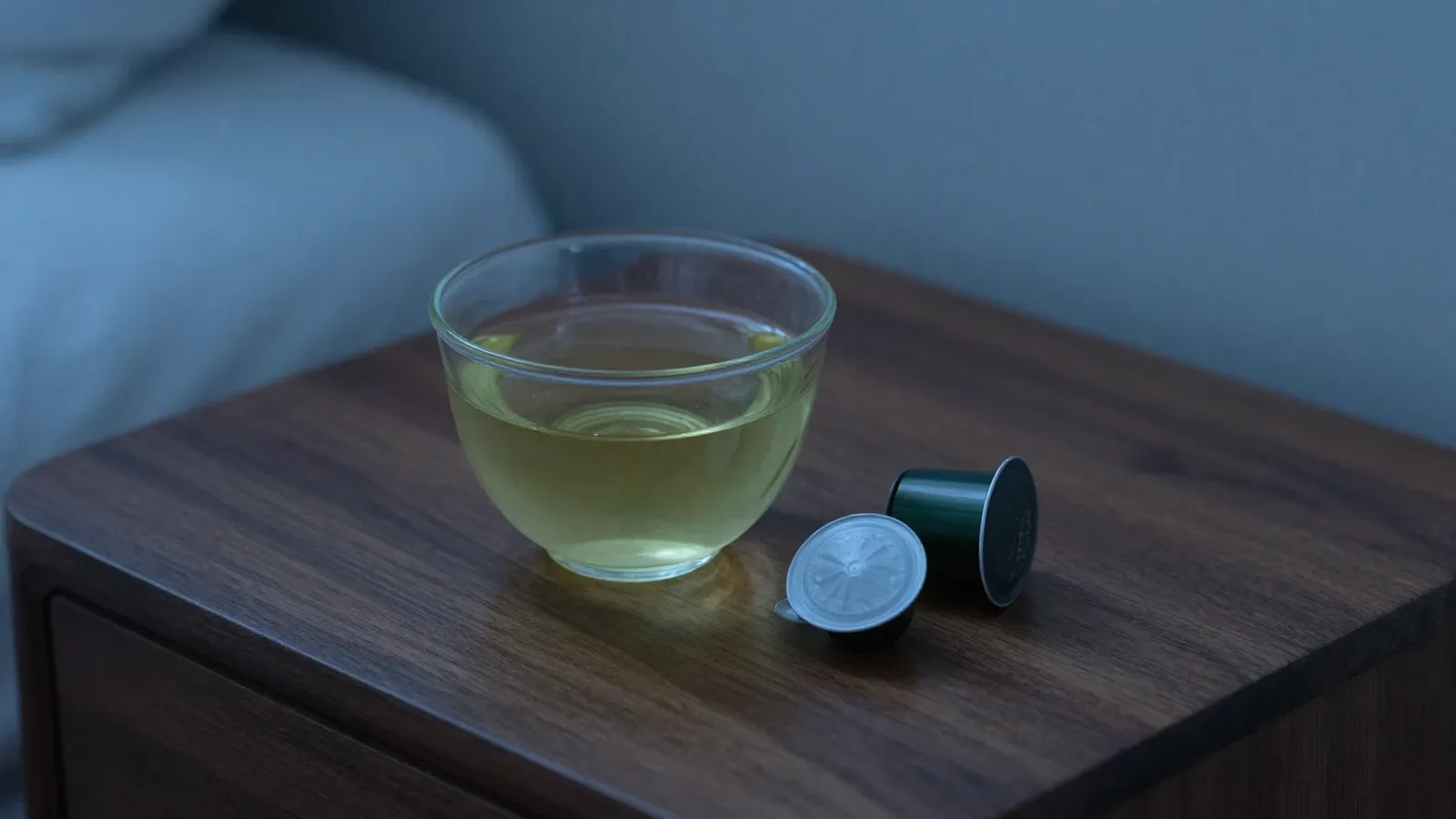 A cup of green tea and a few capsules on a dark nightstand in dim blue evening light.