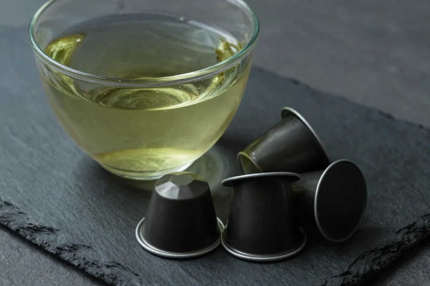 A cup of green tea beside a few capsules on a dark slate surface in soft light.