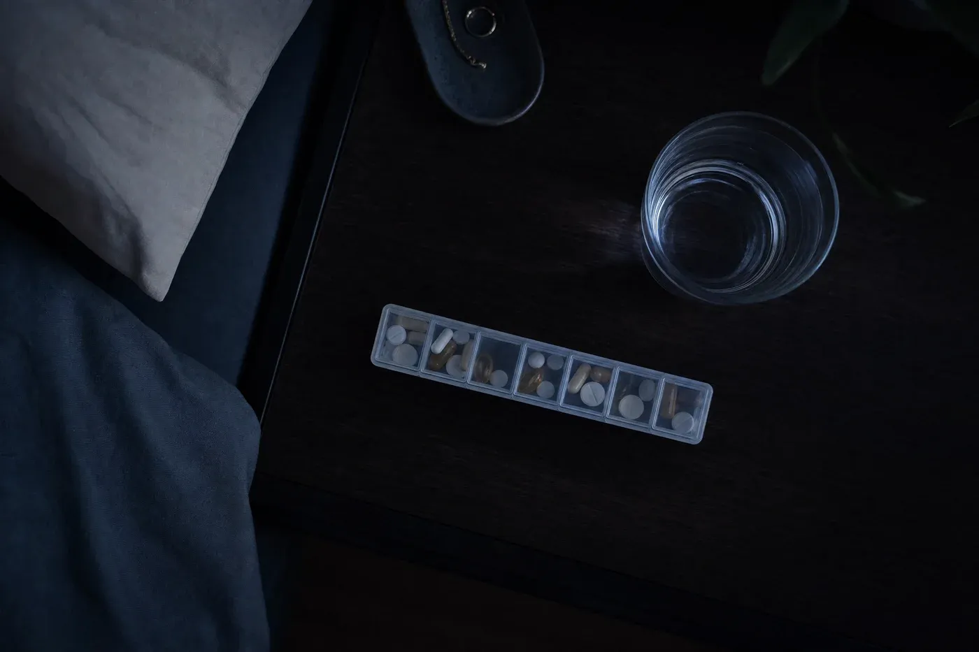 A weekly pill organizer and a glass of water on a bedside table in dim evening light.