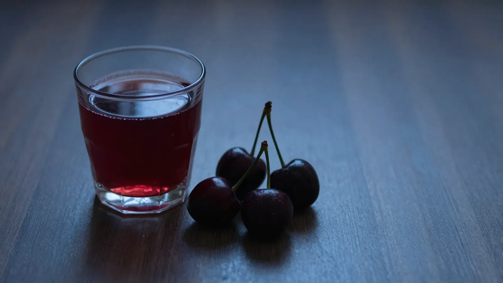 A small glass of dark red juice and a few cherries on a dark surface in dim evening light.
