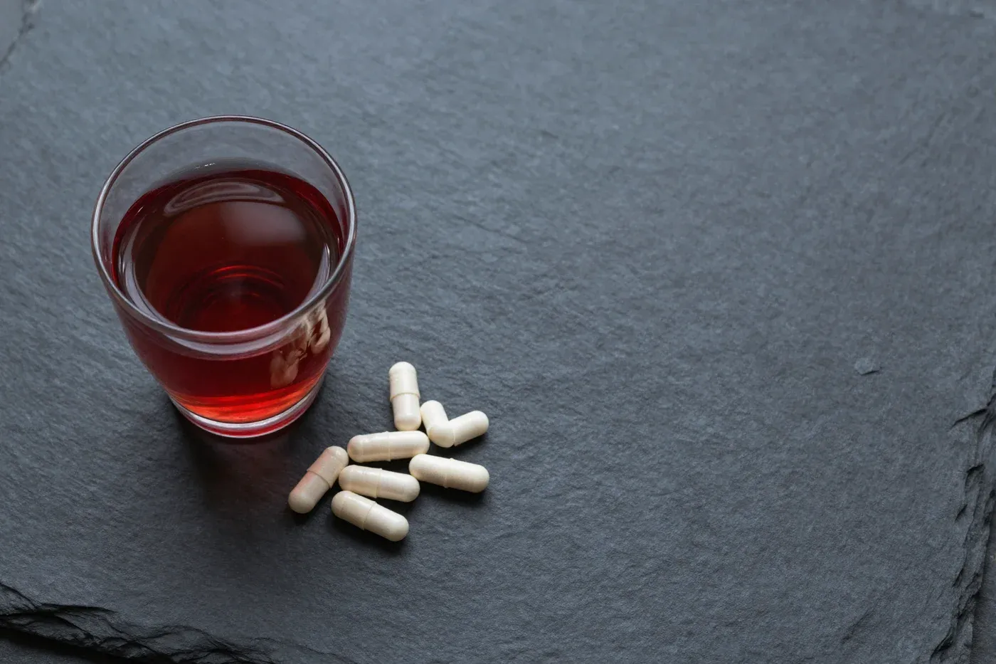 A small glass of dark red juice next to a few capsules on a dark slate surface in soft light.